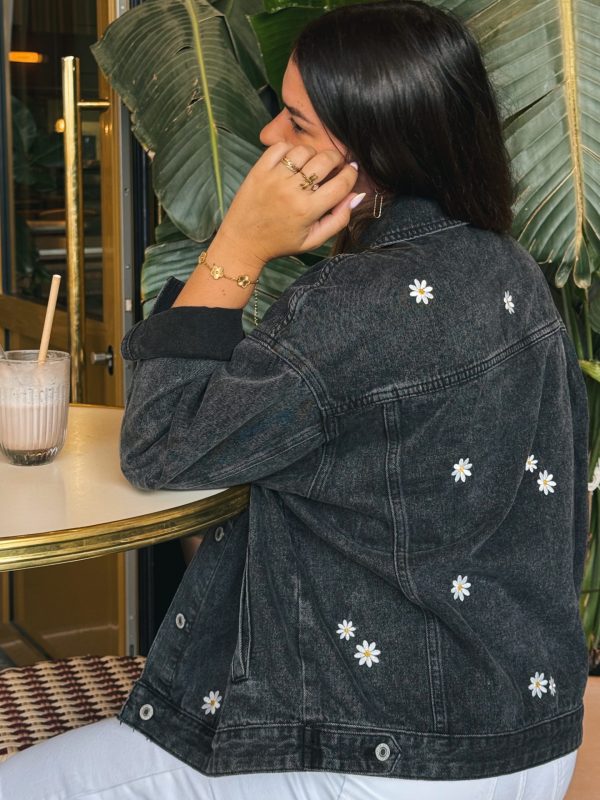 Femme portant une veste en jean avec de la broderie motifs marguerites, sur un tabouret dans un bar